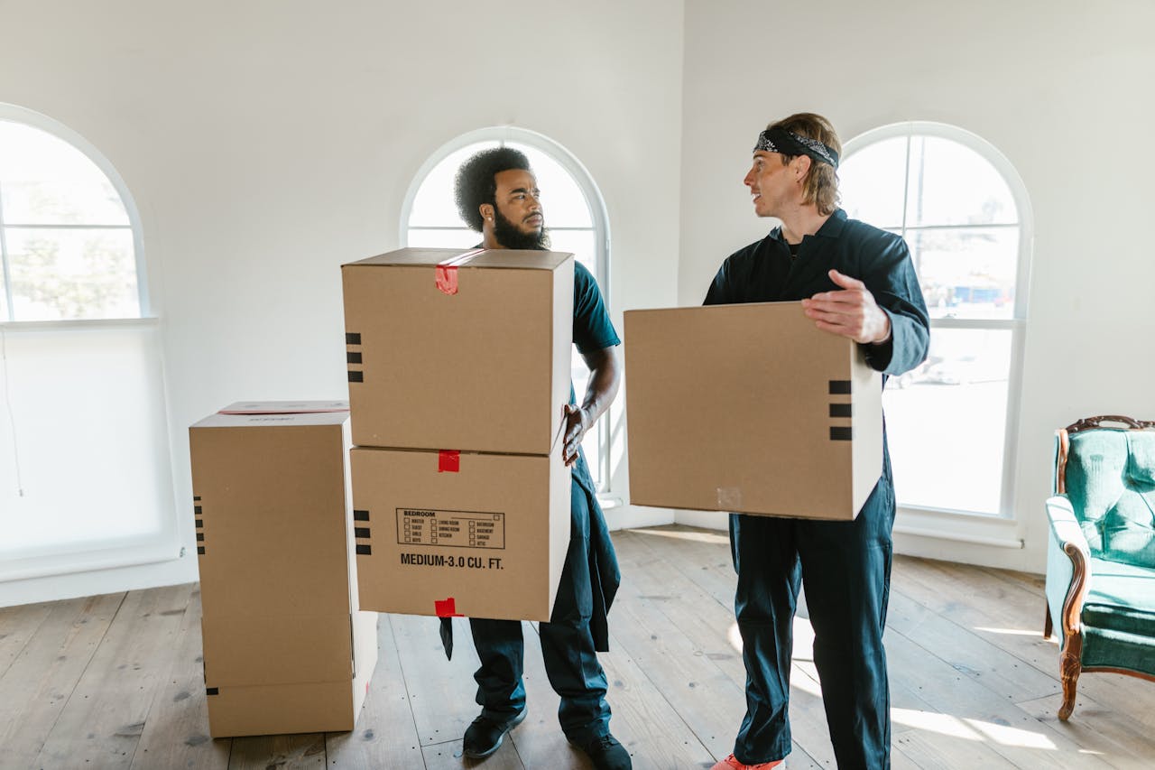 services-06 Two professional movers carrying cardboard boxes inside a bright room with large windows.